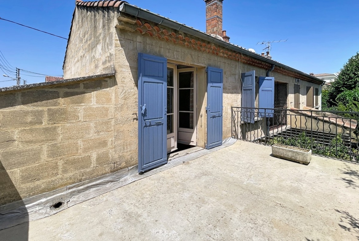 Grande terrasse sur toiture en béton brut avec garde-corps en ferronnerie noire, donnant accès à une façade en pierre de taille avec volets bleus et cheminée en briques rouges. La façade présente deux ouvertures avec volets bleus assortis sous un toit à faible pente recouvert de tuiles canal.