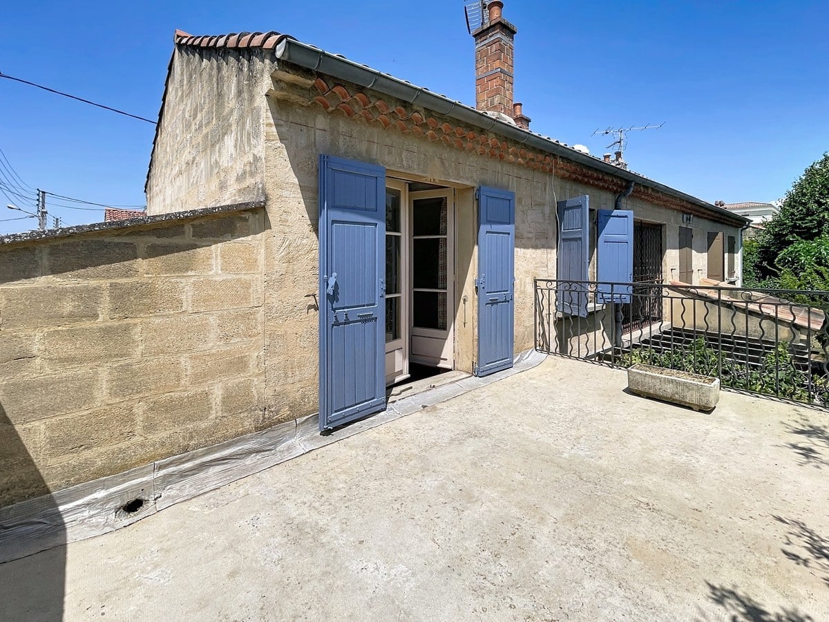 Grande terrasse sur toiture en béton brut avec garde-corps en ferronnerie noire, donnant accès à une façade en pierre de taille avec volets bleus et cheminée en briques rouges. La façade présente deux ouvertures avec volets bleus assortis sous un toit à faible pente recouvert de tuiles canal.
