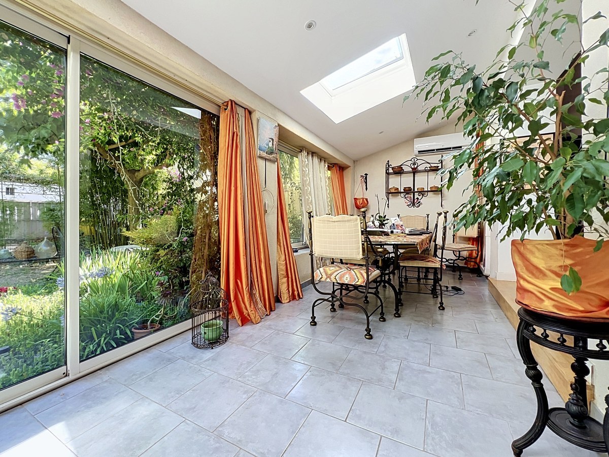 Véranda avec sol en carrelage gris clair, grande baie vitrée coulissante ouvrant sur un jardin arboré et puits de lumière vitré en toiture. Table de salle à manger en fer forgé entourée de chaises assorties, rideaux orangés, climatisation murale et ficus en pot décoratif au premier plan.