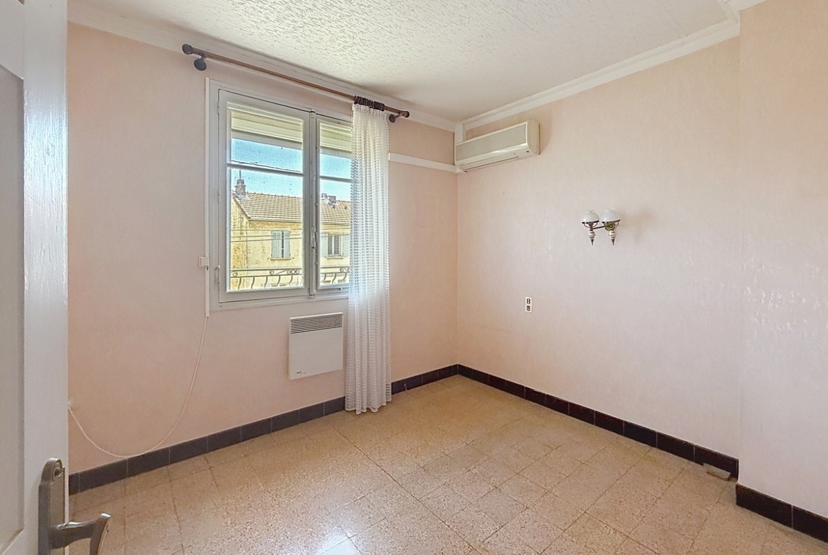 Chambre vide aux murs peints en rose pâle, équipée d'un radiateur électrique sous la fenêtre à double vantail et d'une unité de climatisation réversible en applique murale. Le sol est recouvert d'un carrelage beige granito, une applique murale décorative à globe blanc est fixée sur le mur latéral, et une moulure en plâtre court en périphérie du plafond blanc texturé.
