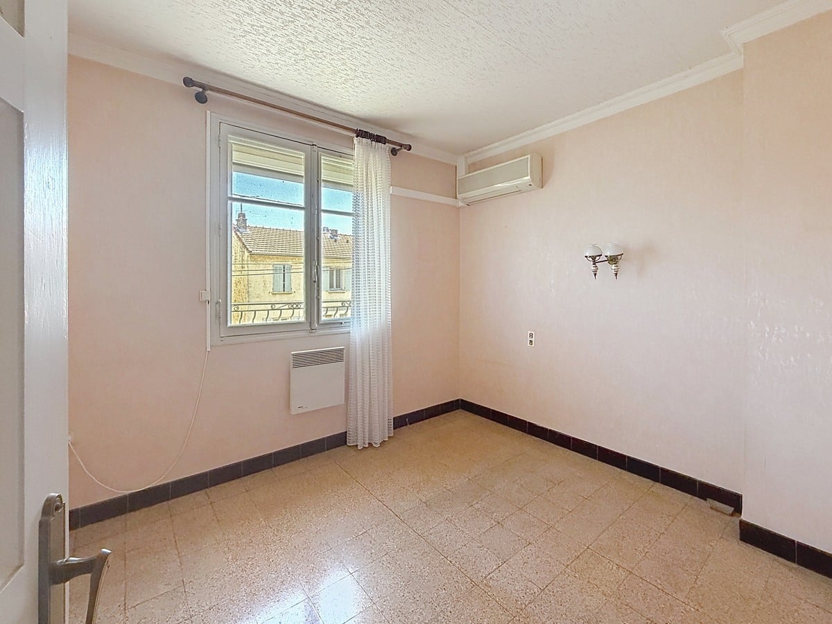 Chambre vide aux murs peints en rose pâle, équipée d'un radiateur électrique sous la fenêtre à double vantail et d'une unité de climatisation réversible en applique murale. Le sol est recouvert d'un carrelage beige granito, une applique murale décorative à globe blanc est fixée sur le mur latéral, et une moulure en plâtre court en périphérie du plafond blanc texturé.