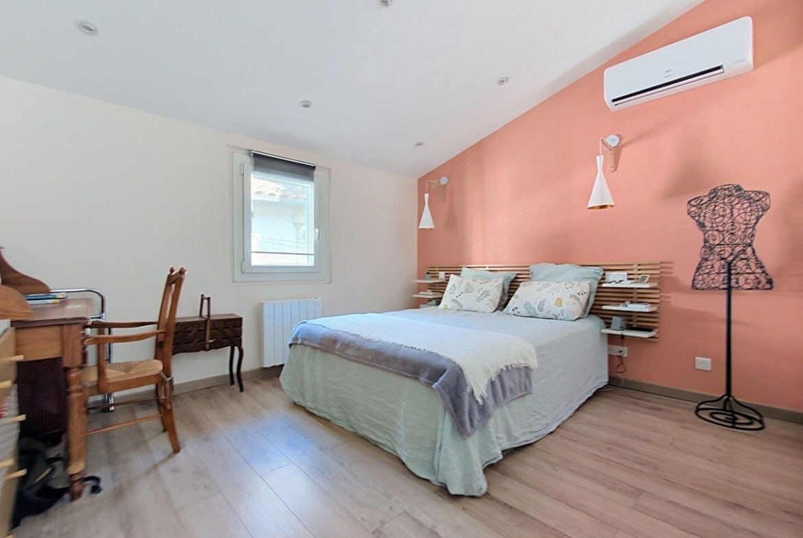 Chambre avec parquet stratifié clair et mur de tête de lit peint en rose terracotta, équipée d'un lit double avec tête de lit en lattes de bois naturel, de deux appliques orientables blanches et d'un climatiseur mural blanc. Un bureau en bois foncé avec chaise cannée ancienne est disposé contre le mur gauche, et un mannequin de couture en fil métallique noir occupe l'angle droit de la pièce.