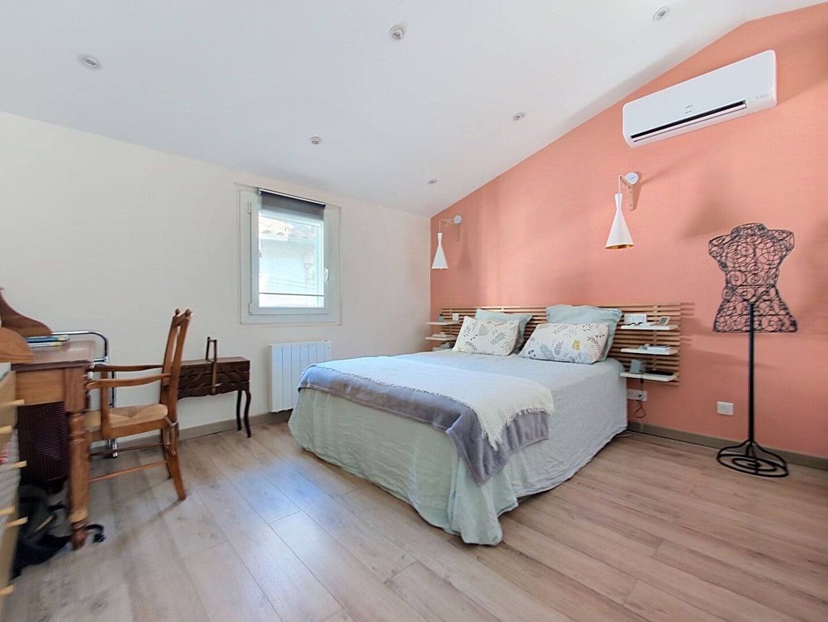 Chambre avec parquet stratifié clair et mur de tête de lit peint en rose terracotta, équipée d'un lit double avec tête de lit en lattes de bois naturel, de deux appliques orientables blanches et d'un climatiseur mural blanc. Un bureau en bois foncé avec chaise cannée ancienne est disposé contre le mur gauche, et un mannequin de couture en fil métallique noir occupe l'angle droit de la pièce.