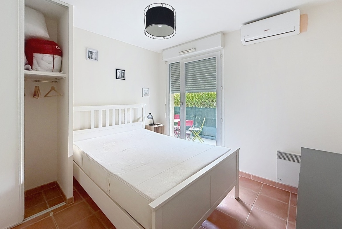 Chambre aux murs blancs avec un lit double à tête de lit blanche en bois, dotée d'un placard encastré ouvert sur la gauche avec étagères et penderie. La pièce est équipée d'une unité de climatisation murale, d'un radiateur électrique en bas à droite, et d'une porte-fenêtre à volet roulant donnant directement sur la terrasse extérieure avec les chaises colorées visibles depuis la chambre.