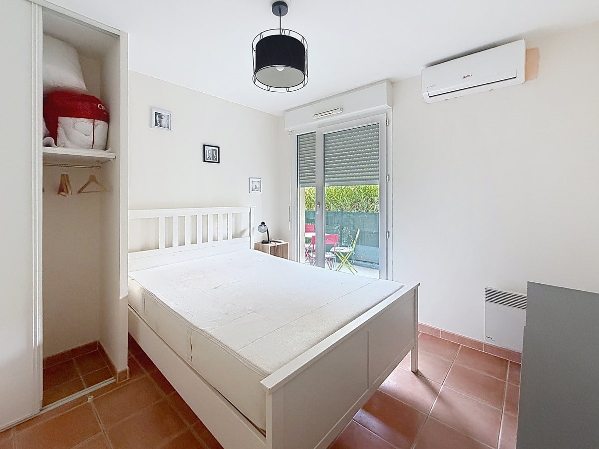 Chambre aux murs blancs avec un lit double à tête de lit blanche en bois, dotée d'un placard encastré ouvert sur la gauche avec étagères et penderie. La pièce est équipée d'une unité de climatisation murale, d'un radiateur électrique en bas à droite, et d'une porte-fenêtre à volet roulant donnant directement sur la terrasse extérieure avec les chaises colorées visibles depuis la chambre.