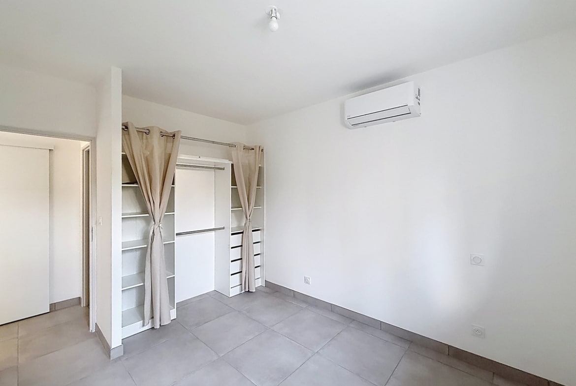 Chambre à carrelage gris clair avec dressing encastré composé de penderies, étagères et tiroirs blancs, dissimulé par des rideaux à œillets en lin beige. Un climatiseur mural blanc est fixé en hauteur sur le mur latéral, et une porte coulissante blanche est visible en fond de pièce.