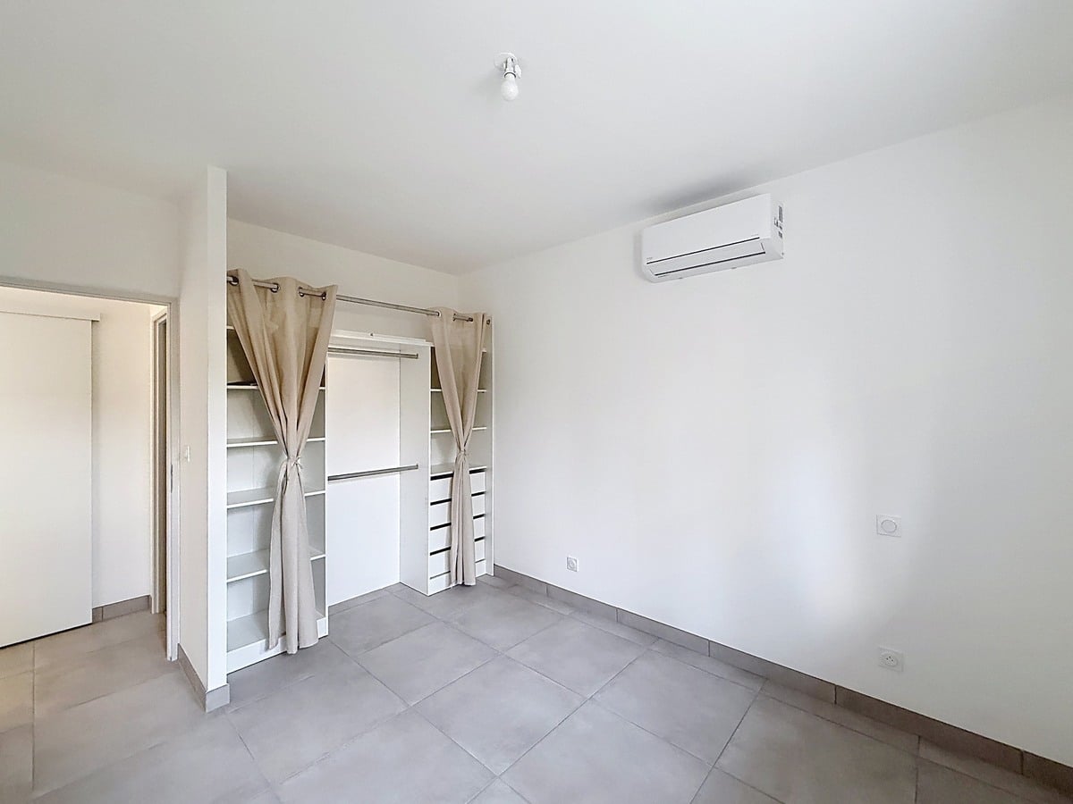 Chambre à carrelage gris clair avec dressing encastré composé de penderies, étagères et tiroirs blancs, dissimulé par des rideaux à œillets en lin beige. Un climatiseur mural blanc est fixé en hauteur sur le mur latéral, et une porte coulissante blanche est visible en fond de pièce.