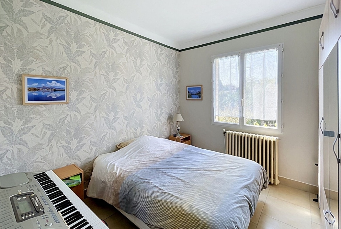 Chambre double avec papier peint à motifs végétaux gris et blanc, lit double avec couverture beige et grise, radiateur en fonte blanc sous la fenêtre PVC à double vitrage donnant sur la végétation extérieure. Clavier électronique numérique posé au pied du lit, armoire blanche encastrée sur la droite, sol en carrelage beige.