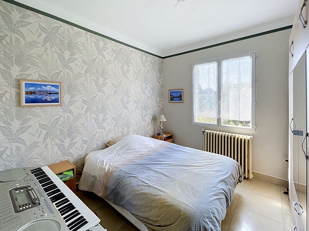 Chambre double avec papier peint à motifs végétaux gris et blanc, lit double avec couverture beige et grise, radiateur en fonte blanc sous la fenêtre PVC à double vitrage donnant sur la végétation extérieure. Clavier électronique numérique posé au pied du lit, armoire blanche encastrée sur la droite, sol en carrelage beige.