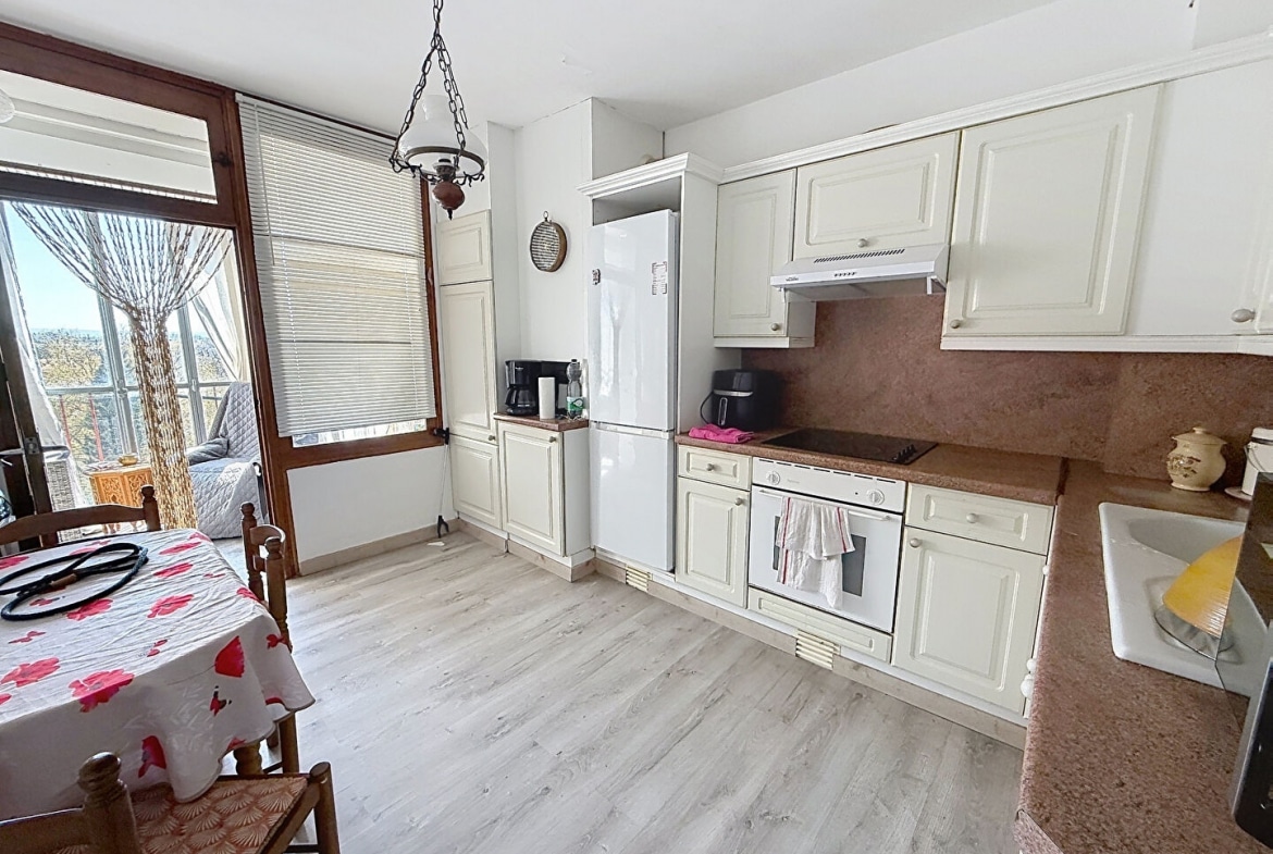 Cuisine équipée avec façades de meubles blanc cassé de style classique, plan de travail et crédence en pierre rose, comprenant une plaque de cuisson encastrée, un four intégré et un grand réfrigérateur blanc. Une table en bois avec deux chaises paillées occupe l'espace repas, face à une porte-fenêtre en bois donnant accès à la loggia.