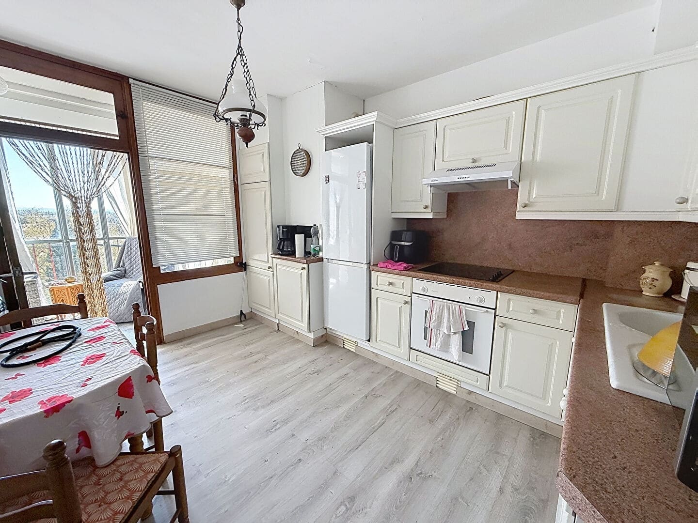 Cuisine équipée avec façades de meubles blanc cassé de style classique, plan de travail et crédence en pierre rose, comprenant une plaque de cuisson encastrée, un four intégré et un grand réfrigérateur blanc. Une table en bois avec deux chaises paillées occupe l'espace repas, face à une porte-fenêtre en bois donnant accès à la loggia.