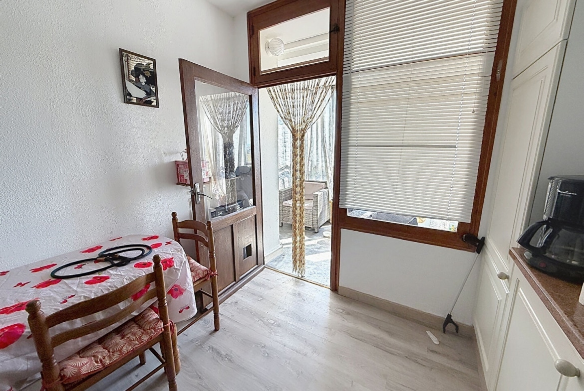 Vue depuis la cuisine vers la porte-fenêtre en bois ouverte donnant sur la loggia, avec un rideau de perles en bois tressé en décoration. L'espace repas comprend une petite table recouverte d'une nappe blanche à motifs rouges et deux chaises en bois à assise paillée, avec des meubles de cuisine blancs visibles à droite.