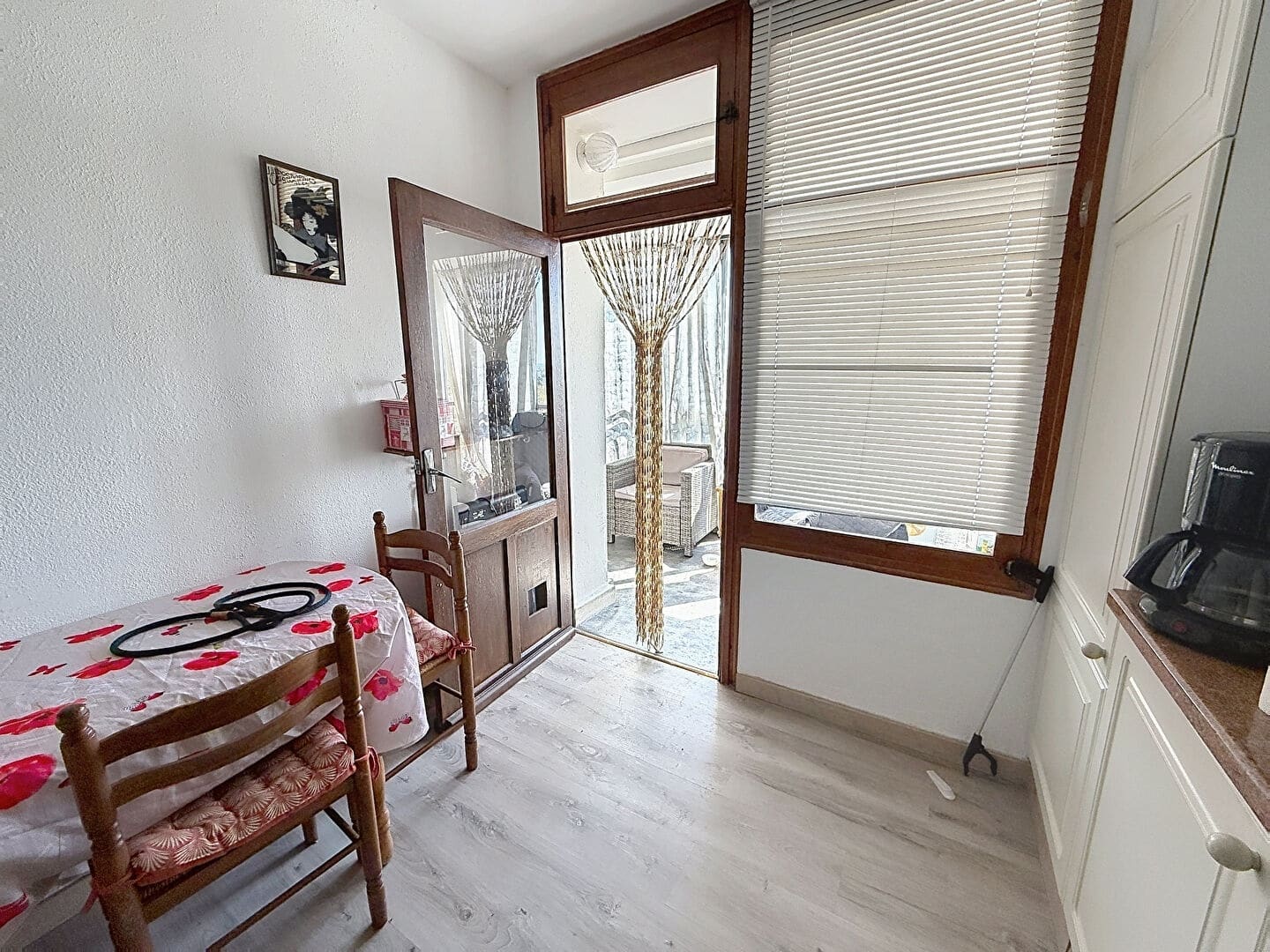 Vue depuis la cuisine vers la porte-fenêtre en bois ouverte donnant sur la loggia, avec un rideau de perles en bois tressé en décoration. L'espace repas comprend une petite table recouverte d'une nappe blanche à motifs rouges et deux chaises en bois à assise paillée, avec des meubles de cuisine blancs visibles à droite.