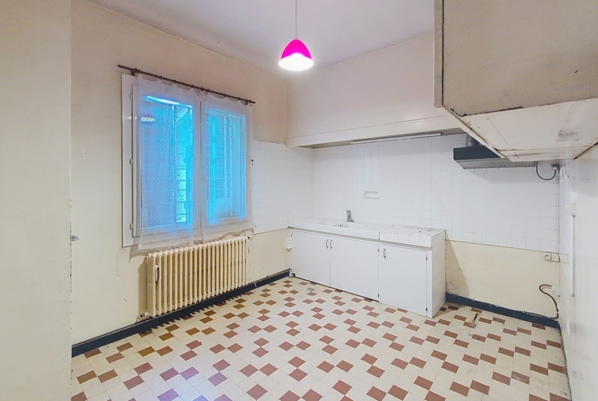 Cuisine vide équipée d'un évier encastré dans un meuble bas blanc, d'un radiateur en fonte crème sous une fenêtre à rideaux blancs et d'une hotte murale blanche. Le sol est recouvert d'un carrelage en damier blanc, bordeaux et ocre, et une suspension rose fuchsia est accrochée au plafond.
