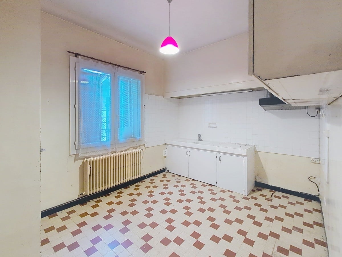 Cuisine vide équipée d'un évier encastré dans un meuble bas blanc, d'un radiateur en fonte crème sous une fenêtre à rideaux blancs et d'une hotte murale blanche. Le sol est recouvert d'un carrelage en damier blanc, bordeaux et ocre, et une suspension rose fuchsia est accrochée au plafond.