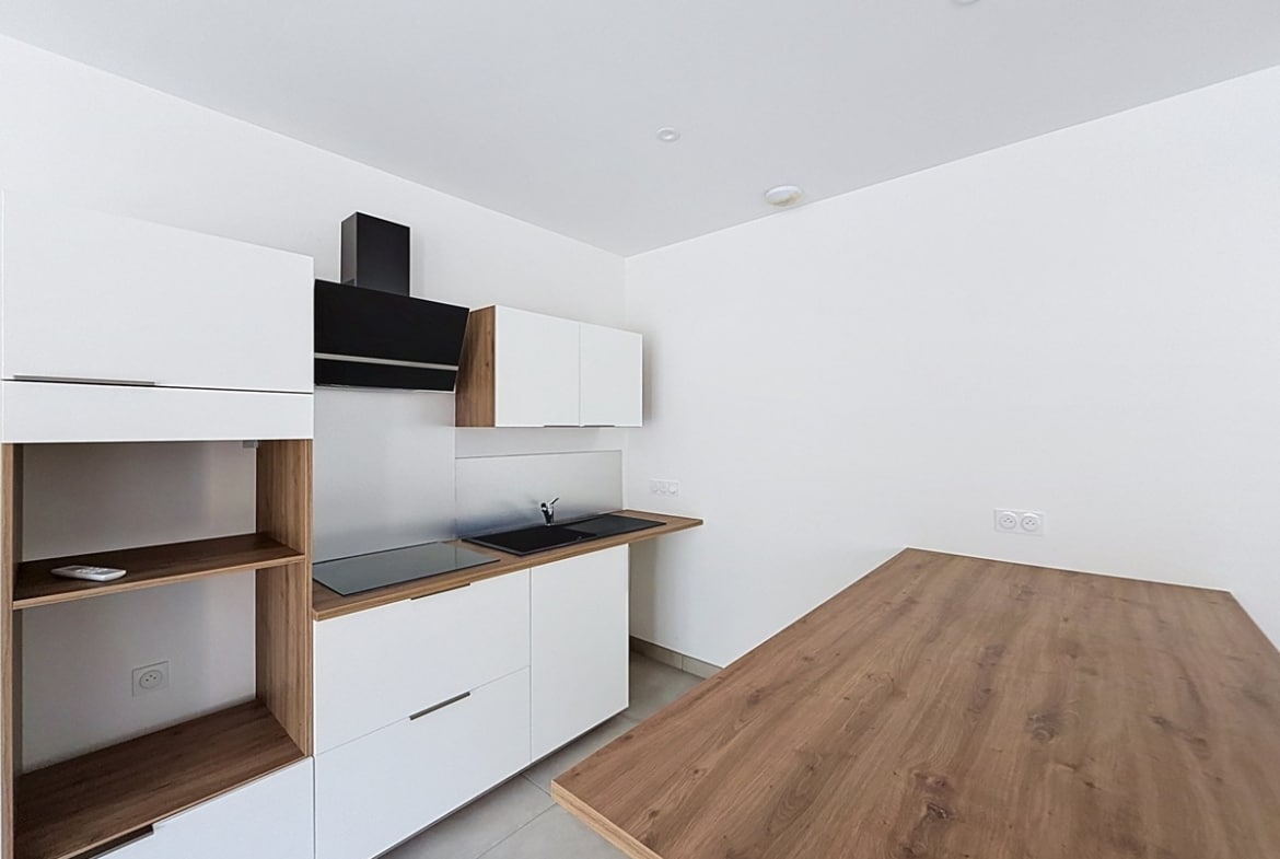 Cuisine linéaire avec façades blanches mates et plan de travail bois, intégrant une plaque de cuisson noire encastrée, un évier noir et une hotte murale noire, complétée par des éléments de rangement ouverts en bois naturel. Un îlot en bois clair occupe le premier plan face à la cuisine, sur un carrelage gris clair.