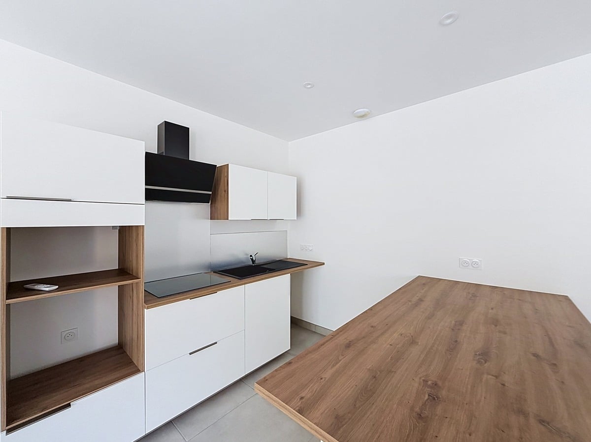 Cuisine linéaire avec façades blanches mates et plan de travail bois, intégrant une plaque de cuisson noire encastrée, un évier noir et une hotte murale noire, complétée par des éléments de rangement ouverts en bois naturel. Un îlot en bois clair occupe le premier plan face à la cuisine, sur un carrelage gris clair.