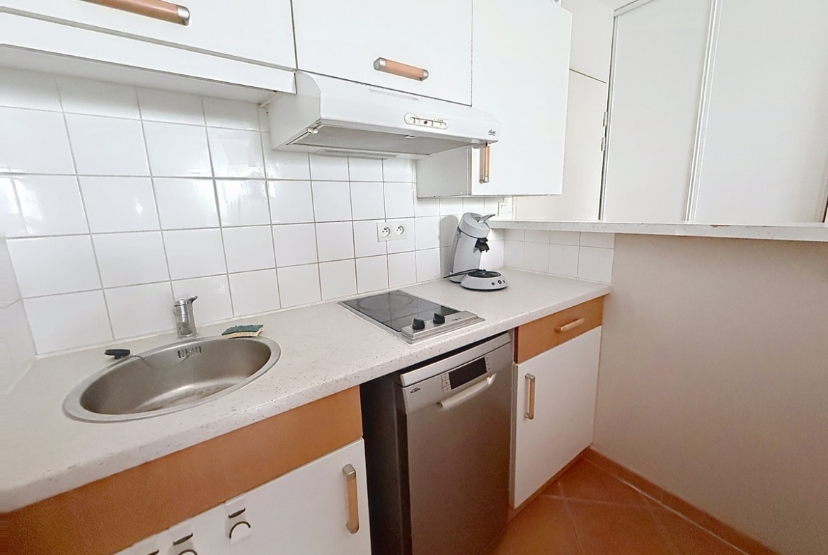 Cuisine en L aux façades blanches et poignées en bois beige, équipée de deux plaques vitrocéramiques, d'un évier rond en inox, d'un lave-vaisselle encastré inox et d'une hotte aspirante murale. Le crédence est entièrement carrelée en blanc, le plan de travail est en pierre reconstituée gris clair, et le sol reprend les carreaux en terre cuite présents dans tout l'appartement.