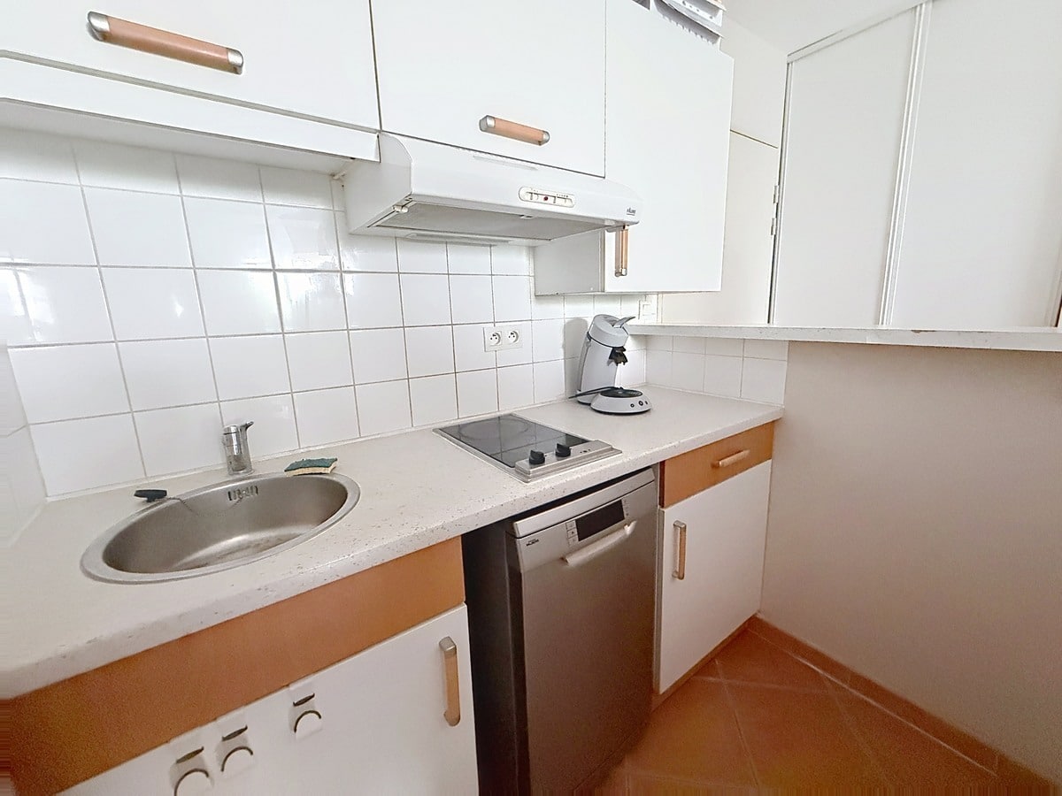Cuisine en L aux façades blanches et poignées en bois beige, équipée de deux plaques vitrocéramiques, d'un évier rond en inox, d'un lave-vaisselle encastré inox et d'une hotte aspirante murale. Le crédence est entièrement carrelée en blanc, le plan de travail est en pierre reconstituée gris clair, et le sol reprend les carreaux en terre cuite présents dans tout l'appartement.