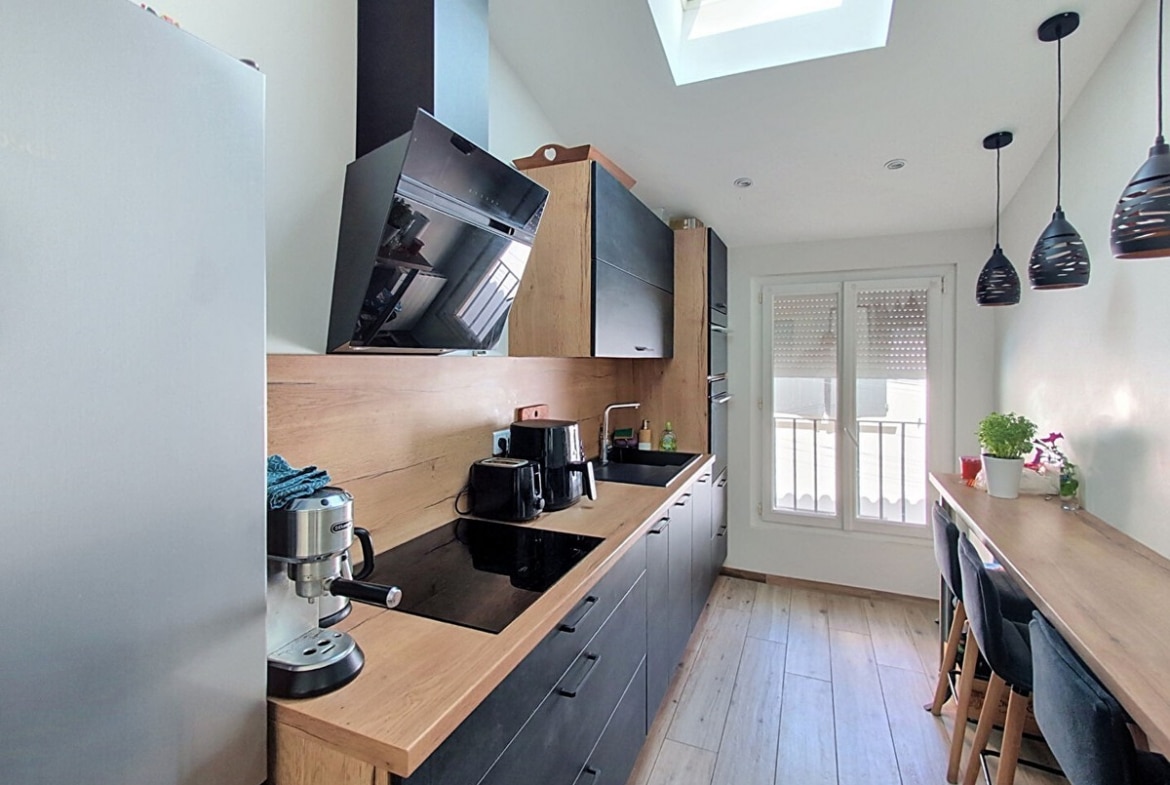 Cuisine linéaire entièrement équipée avec façades gris anthracite et plan de travail en bois clair, dotée d'une hotte noire inclinée et d'une plaque de cuisson à induction, éclairée par un velux en plafond. Un bar en bois avec tabourets gris anthracite et deux suspensions noires en métal ajouré longe le mur opposé côté fenêtre.