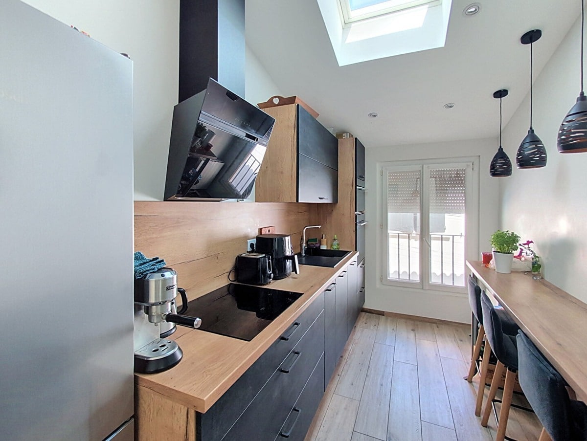 Cuisine linéaire entièrement équipée avec façades gris anthracite et plan de travail en bois clair, dotée d'une hotte noire inclinée et d'une plaque de cuisson à induction, éclairée par un velux en plafond. Un bar en bois avec tabourets gris anthracite et deux suspensions noires en métal ajouré longe le mur opposé côté fenêtre.