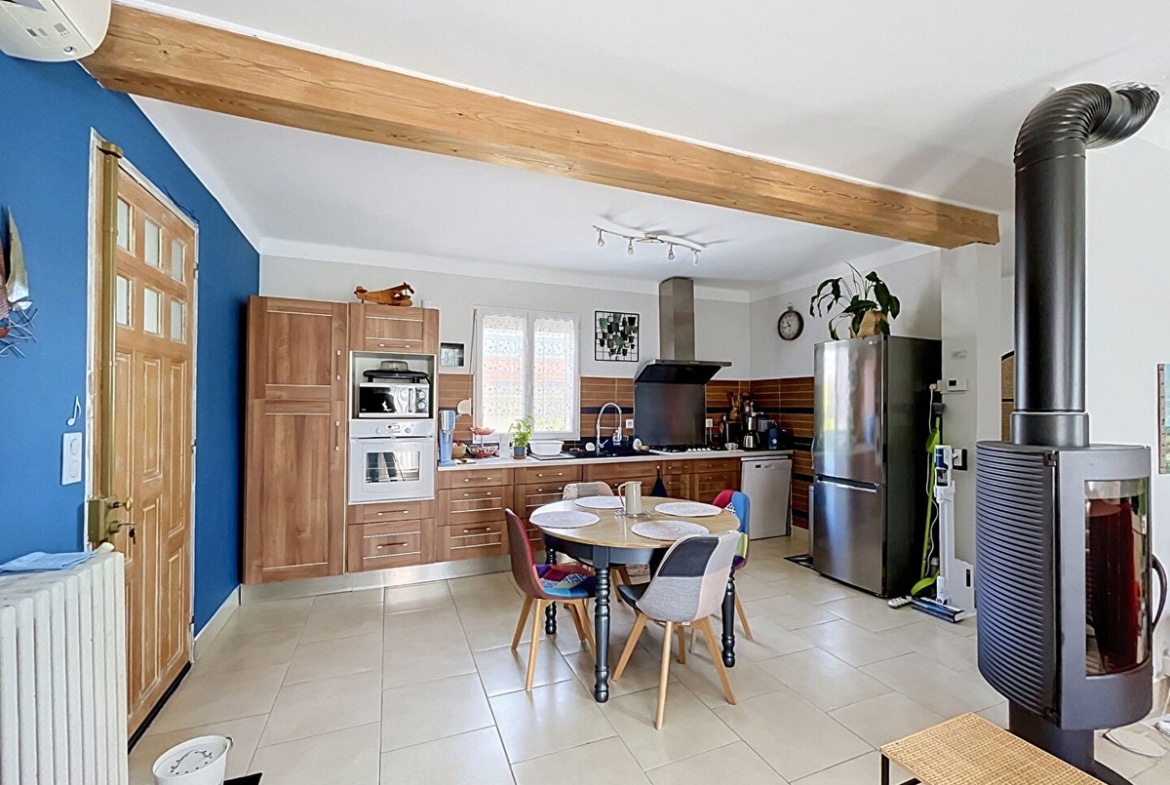 Cuisine ouverte équipée avec façades en bois noyer, four encastré, micro-ondes et réfrigérateur inox, séparée de la salle à manger par un poêle à bois noir sur pied. Table ronde en bois avec quatre chaises dépareillées colorées, mur d'accent bleu cobalt et poutre en bois apparent au plafond.