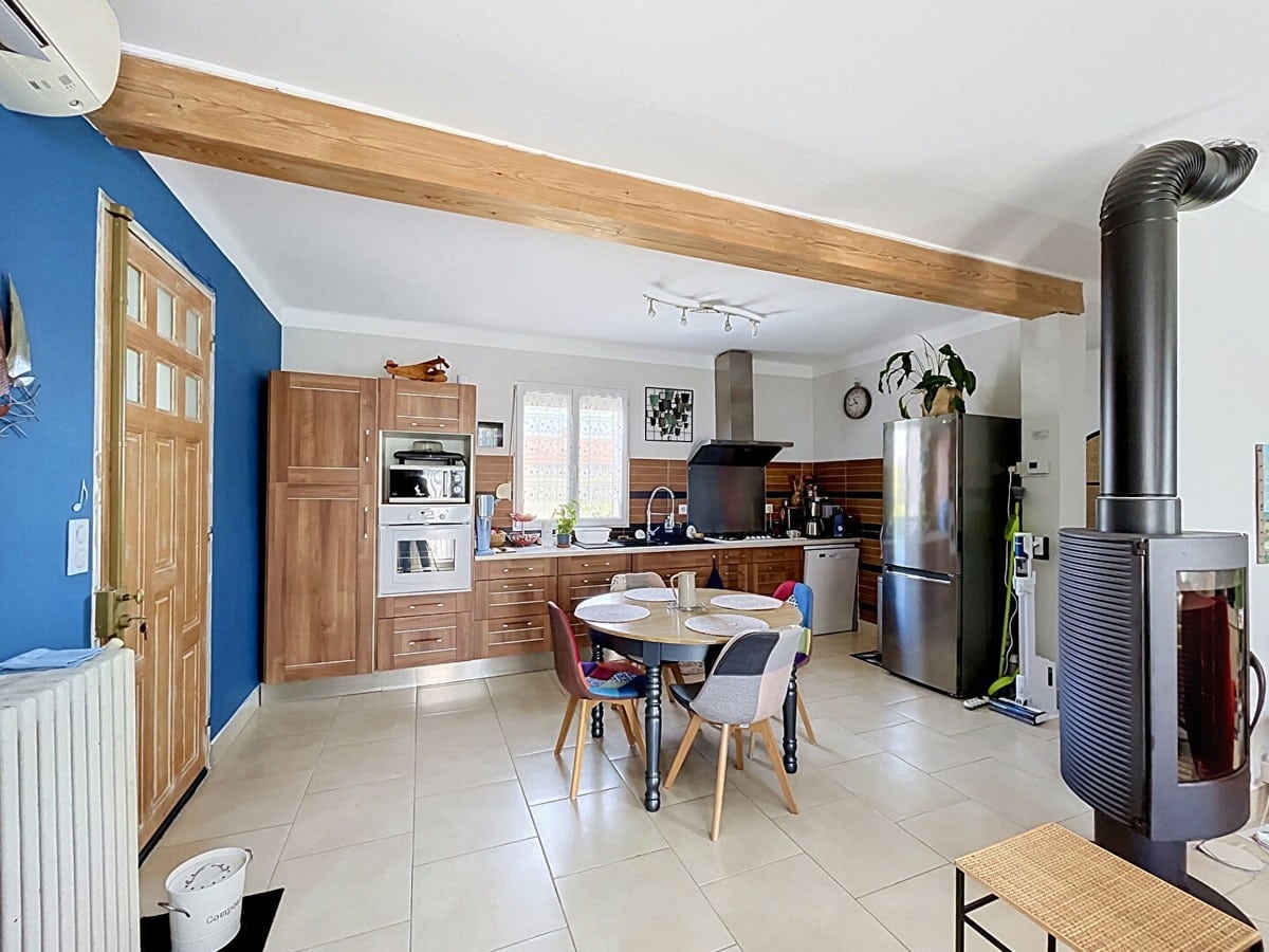 Cuisine ouverte équipée avec façades en bois noyer, four encastré, micro-ondes et réfrigérateur inox, séparée de la salle à manger par un poêle à bois noir sur pied. Table ronde en bois avec quatre chaises dépareillées colorées, mur d'accent bleu cobalt et poutre en bois apparent au plafond.