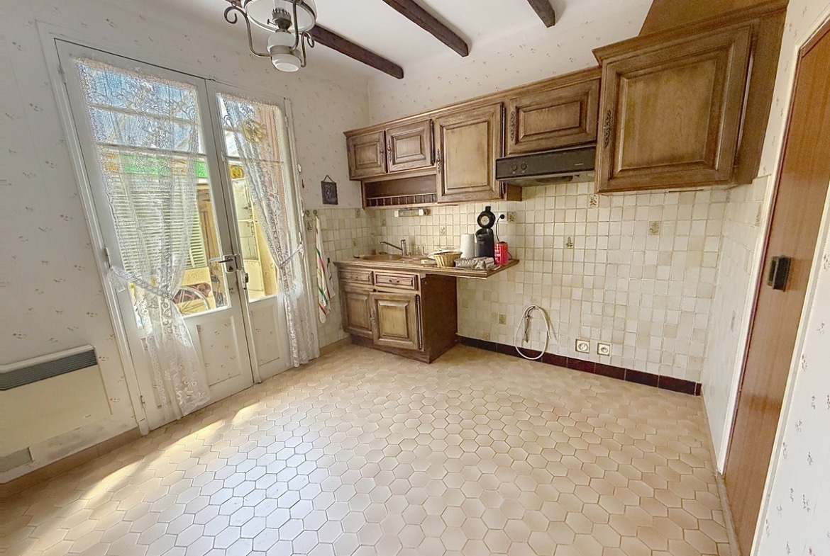 Cuisine indépendante dotée de meubles en bois massif teinte chêne rustique, d'un carrelage mural en faïence blanche petit format et d'un sol en tomettes hexagonales beiges. Le plafond présente des poutres en bois apparentes, une porte-fenêtre à deux vantaux avec rideaux en dentelle donne sur l'extérieur, et un espace pour lave-linge est prévu avec arrivée d'eau visible.