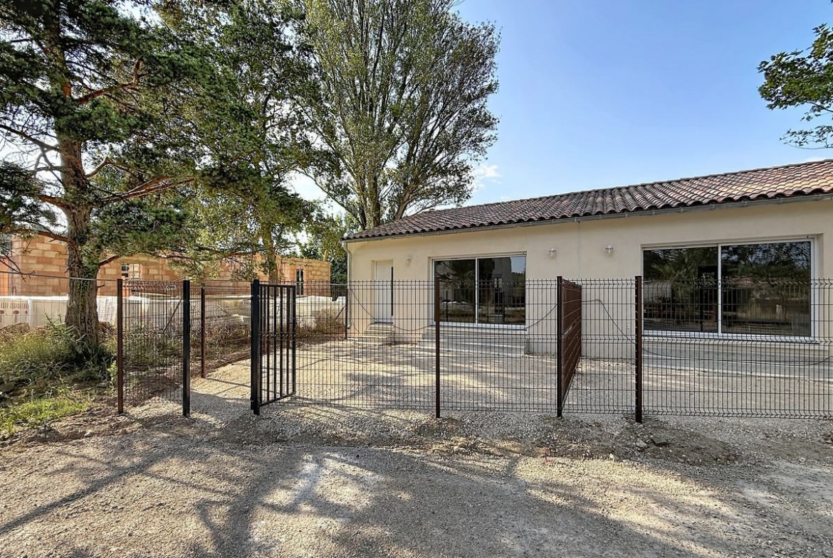 Maison plain-pied à enduit beige clair avec toiture en tuiles canal, entourée d'une clôture grillagée anthracite équipée d'un portillon métallique. La cour avant est revêtue de graviers, avec de grands arbres matures visibles en arrière-plan et une construction en briques adjacente.