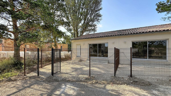 Maison plain-pied à enduit beige clair avec toiture en tuiles canal, entourée d'une clôture grillagée anthracite équipée d'un portillon métallique. La cour avant est revêtue de graviers, avec de grands arbres matures visibles en arrière-plan et une construction en briques adjacente.
