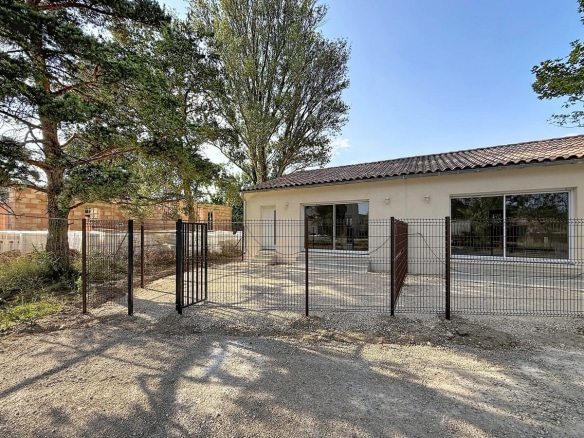 Maison plain-pied à enduit beige clair avec toiture en tuiles canal, entourée d'une clôture grillagée anthracite équipée d'un portillon métallique. La cour avant est revêtue de graviers, avec de grands arbres matures visibles en arrière-plan et une construction en briques adjacente.