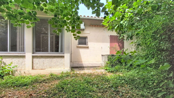 Façade en enduit blanc d'une maison de plain-pied avec toiture en tuiles rondes, grande baie vitrée aluminium et une petite fenêtre avec grilles métalliques grises. Le jardin en façade est envahi de végétation basse et d'arbres à feuilles caduques qui encadrent entièrement la maison.