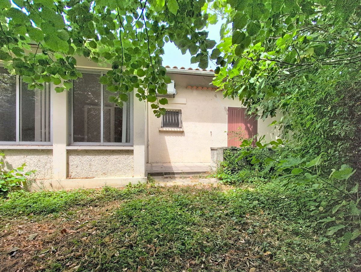 Façade en enduit blanc d'une maison de plain-pied avec toiture en tuiles rondes, grande baie vitrée aluminium et une petite fenêtre avec grilles métalliques grises. Le jardin en façade est envahi de végétation basse et d'arbres à feuilles caduques qui encadrent entièrement la maison.