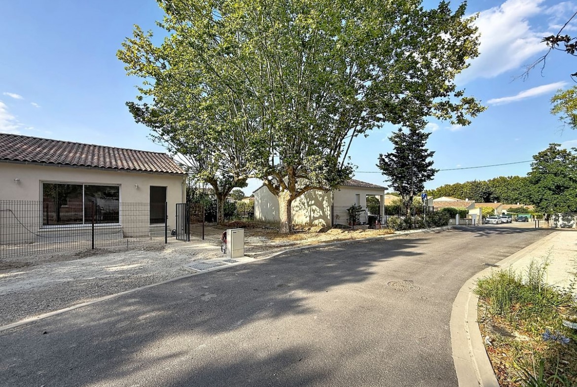 Vue de la rue sur une maison plain-pied à enduit beige avec toiture en tuiles canal et grandes baies vitrées coulissantes, implantée dans un lotissement pavillonaire récent avec voirie asphaltée neuve. Un grand platane et des arbres hauts encadrent la parcelle, avec d'autres maisons neuves similaires visibles en fond de rue.
