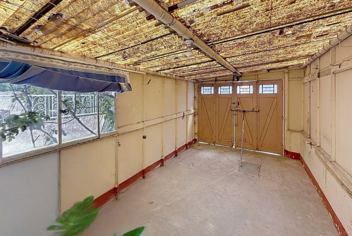 Garage rectangulaire aux murs enduits peints en jaune clair avec une plinthe rouge, fermé par une double porte en bois à panneaux avec impostes vitrées. Le plafond présente une toiture translucide en fibrociment très oxydée reposant sur des rails métalliques apparents, une fenêtre latérale donne sur le jardin et le sol est en béton brut.