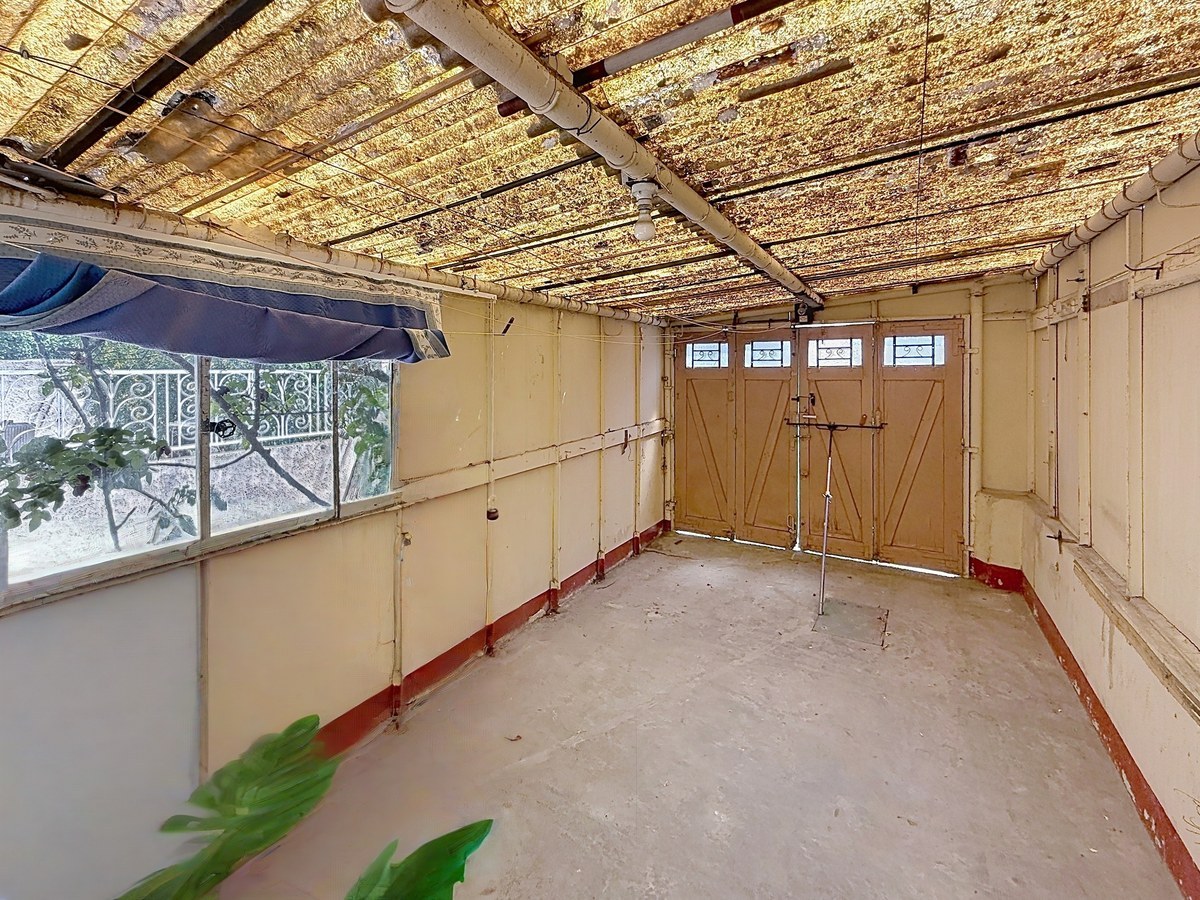 Garage rectangulaire aux murs enduits peints en jaune clair avec une plinthe rouge, fermé par une double porte en bois à panneaux avec impostes vitrées. Le plafond présente une toiture translucide en fibrociment très oxydée reposant sur des rails métalliques apparents, une fenêtre latérale donne sur le jardin et le sol est en béton brut.