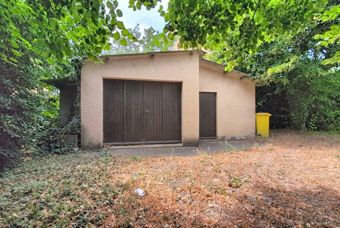 Bâtiment annexe en enduit beige avec une grande double porte en bois sombre et une porte piétonne latérale, entouré d'une végétation dense de grands arbres. Le sol devant le garage est recouvert de feuilles mortes séchées et la poubelle de collecte jaune est visible à droite.