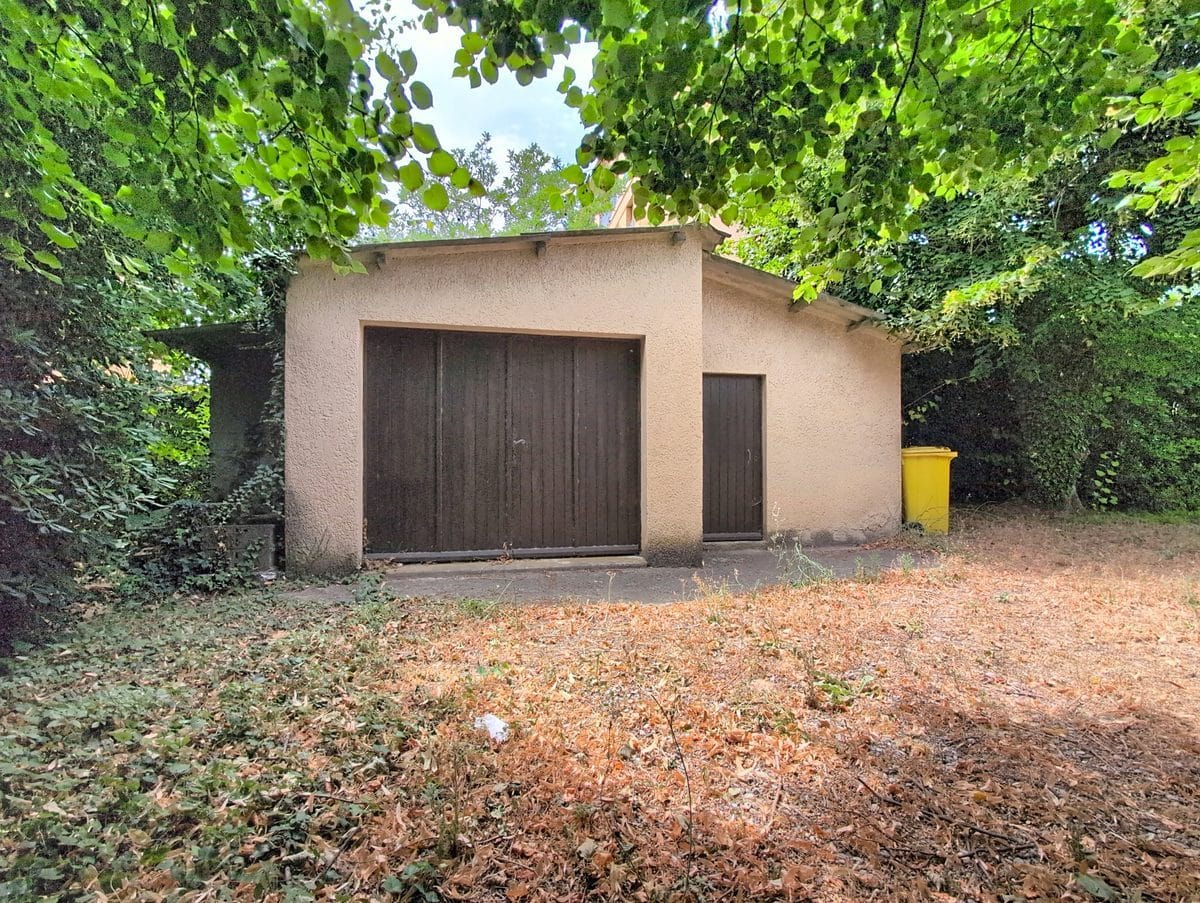Bâtiment annexe en enduit beige avec une grande double porte en bois sombre et une porte piétonne latérale, entouré d'une végétation dense de grands arbres. Le sol devant le garage est recouvert de feuilles mortes séchées et la poubelle de collecte jaune est visible à droite.