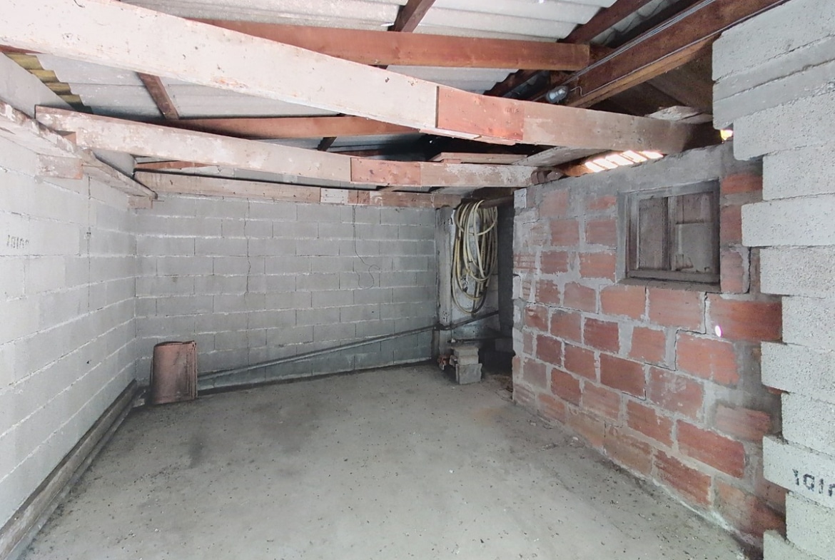 Intérieur de garage en parpaings et briques non enduits, avec sol en béton brut et charpente métallique et bois apparente sous une toiture en fibrociment translucide. Un tuyau souple enroulé est posé dans le fond du local, et un petit soupirail est visible dans le mur en briques rouges sur la droite.