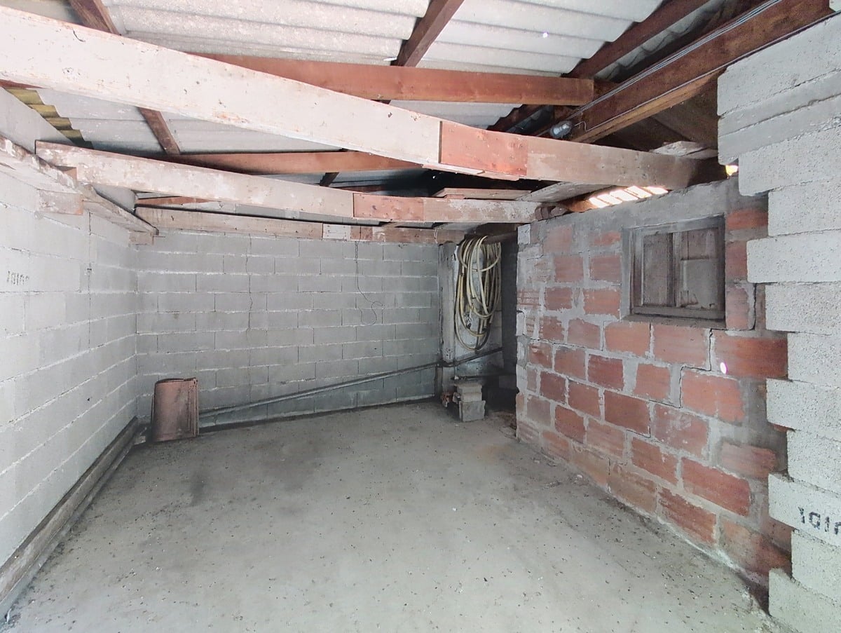 Intérieur de garage en parpaings et briques non enduits, avec sol en béton brut et charpente métallique et bois apparente sous une toiture en fibrociment translucide. Un tuyau souple enroulé est posé dans le fond du local, et un petit soupirail est visible dans le mur en briques rouges sur la droite.