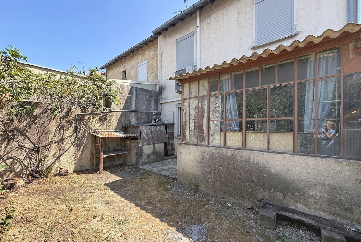Cour arrière délimitée par des murs en béton enduit, avec un espace jardin au sol sec et terreux comportant un arbuste et un cabanon de rangement au toit en fibrociment ondulé. La façade arrière de la maison révèle une véranda ancienne aux châssis métalliques vitrés très dégradés, un volet roulant blanc à l'étage et un climatiseur mural en façade.