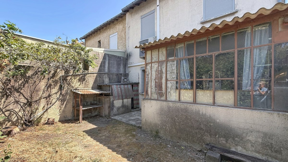 Cour arrière délimitée par des murs en béton enduit, avec un espace jardin au sol sec et terreux comportant un arbuste et un cabanon de rangement au toit en fibrociment ondulé. La façade arrière de la maison révèle une véranda ancienne aux châssis métalliques vitrés très dégradés, un volet roulant blanc à l'étage et un climatiseur mural en façade.