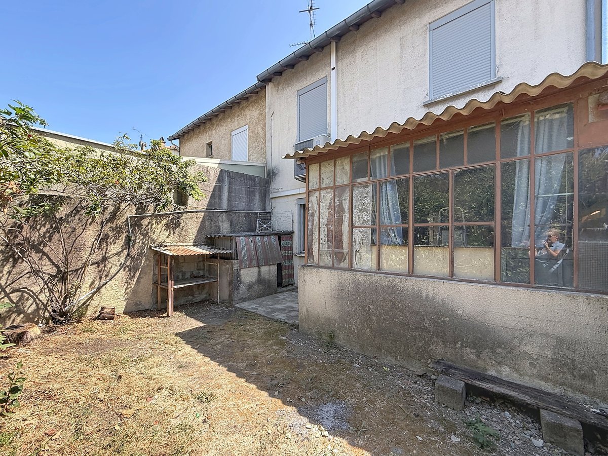 Cour arrière délimitée par des murs en béton enduit, avec un espace jardin au sol sec et terreux comportant un arbuste et un cabanon de rangement au toit en fibrociment ondulé. La façade arrière de la maison révèle une véranda ancienne aux châssis métalliques vitrés très dégradés, un volet roulant blanc à l'étage et un climatiseur mural en façade.