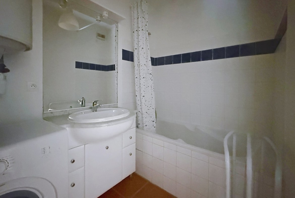 Salle de bain carrelée blanc avec frise de carreaux bleus marine en hauteur, équipée d'une baignoire rectangulaire avec rideau de douche à motifs, d'un meuble vasque blanc à portes avec miroir éclairé au-dessus. Un lave-linge est installé en angle gauche sous un chauffe-eau mural, le sol est revêtu de carreaux en terre cuite rouge.