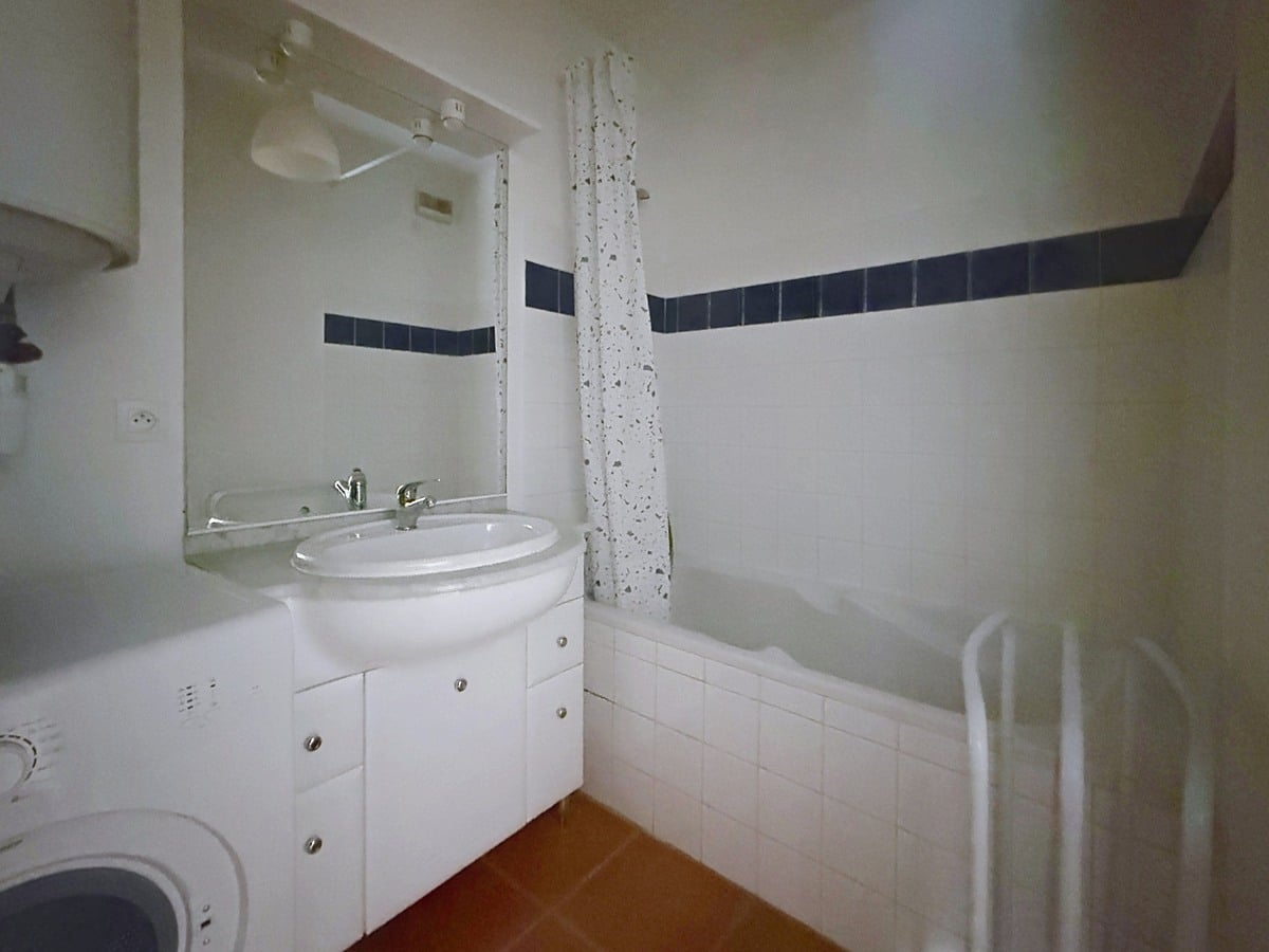 Salle de bain carrelée blanc avec frise de carreaux bleus marine en hauteur, équipée d'une baignoire rectangulaire avec rideau de douche à motifs, d'un meuble vasque blanc à portes avec miroir éclairé au-dessus. Un lave-linge est installé en angle gauche sous un chauffe-eau mural, le sol est revêtu de carreaux en terre cuite rouge.