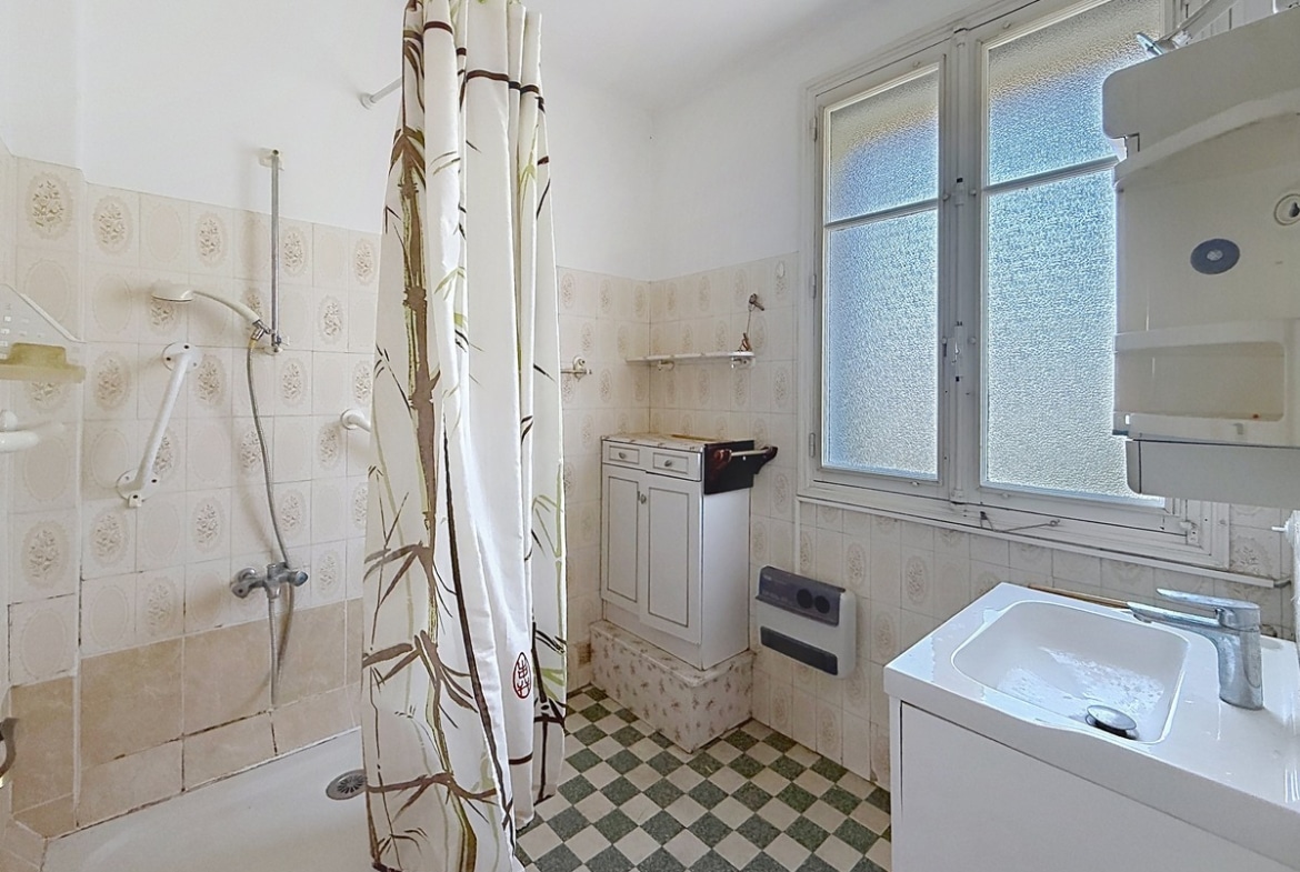 Salle de bain ancienne équipée d'une douche à l'italienne avec rideau à motifs bambou, d'un meuble vasque blanc moderne et d'un petit meuble de rangement sous-lavabo blanc. Le sol présente un carrelage damier vert et blanc, les murs sont revêtus de faïence beige à motifs floraux en relief, et une fenêtre à vitrage dépoli apporte la lumière naturelle.