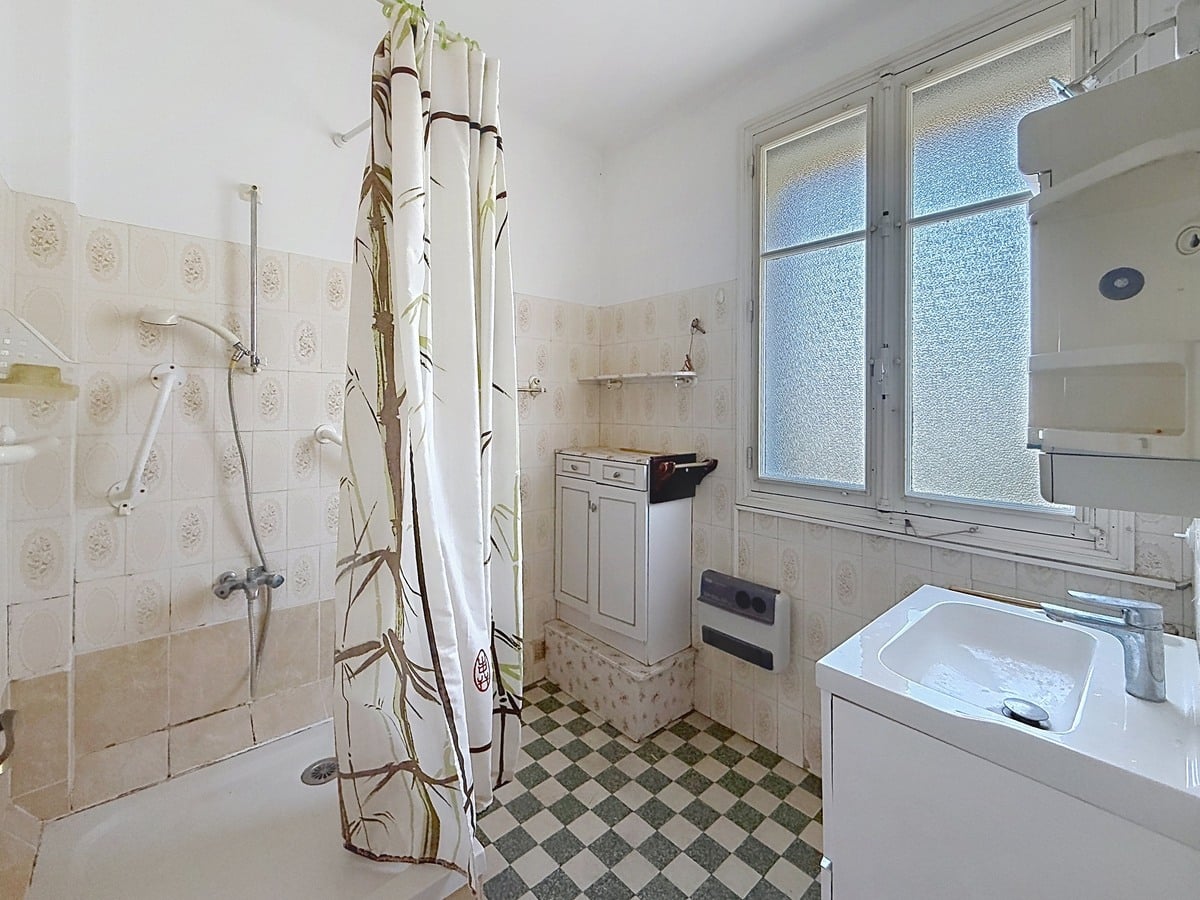 Salle de bain ancienne équipée d'une douche à l'italienne avec rideau à motifs bambou, d'un meuble vasque blanc moderne et d'un petit meuble de rangement sous-lavabo blanc. Le sol présente un carrelage damier vert et blanc, les murs sont revêtus de faïence beige à motifs floraux en relief, et une fenêtre à vitrage dépoli apporte la lumière naturelle.