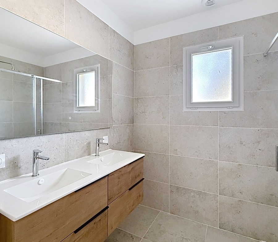 Salle de bain carrelée du sol au plafond avec grand format pierre beige, équipée d'un meuble vasque suspendu en bois clair à double vasque blanche et robinetterie chromée, avec miroir pleine largeur au-dessus. Une douche à l'italienne est partiellement visible à droite, deux fenêtres à verre dépoli assurent la ventilation et la lumière naturelle.