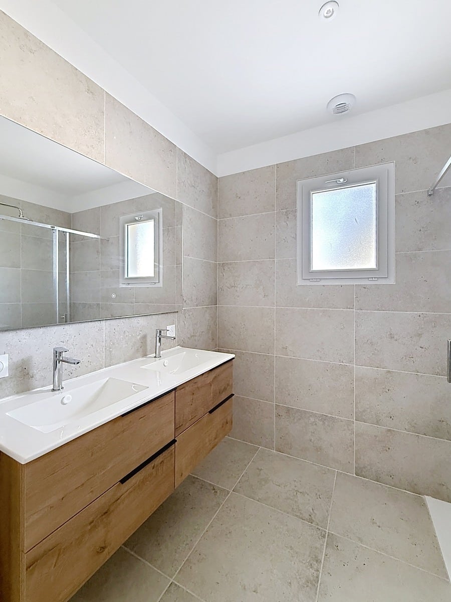 Salle de bain carrelée du sol au plafond avec grand format pierre beige, équipée d'un meuble vasque suspendu en bois clair à double vasque blanche et robinetterie chromée, avec miroir pleine largeur au-dessus. Une douche à l'italienne est partiellement visible à droite, deux fenêtres à verre dépoli assurent la ventilation et la lumière naturelle.
