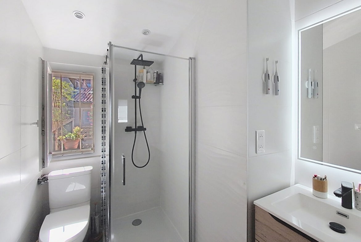 Salle d'eau entièrement carrelée en grand format blanc, équipée d'une douche à l'italienne avec paroi en verre, colonne de douche noire avec pommeau carré et douchette, et d'un meuble vasque suspendu en bois avec plan vasque blanc et miroir LED rectangulaire. Des WC suspendus sont installés contre le mur gauche sous une fenêtre ouvrant sur l'extérieur, avec un décor de mosaïque à reflets chromés en bande verticale.